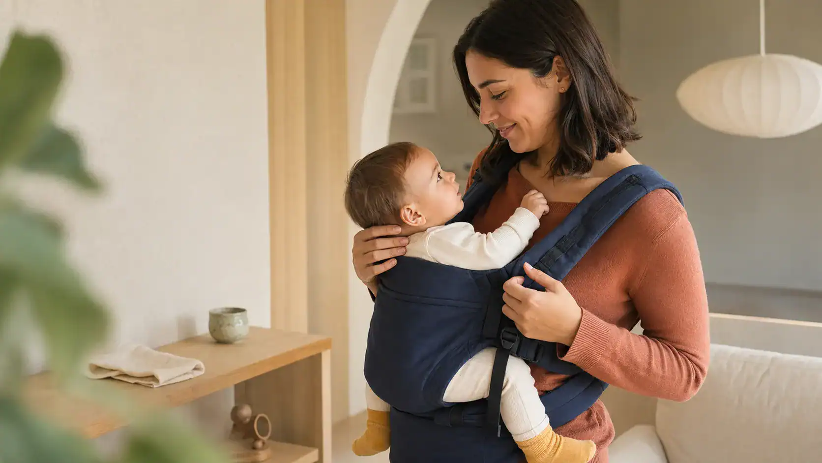 Mutter trägt ein waches Baby in einer Babytrage im Alltagsmoment, warmes natürliches Tageslicht, geborgene Nähe